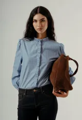 Camisa de manga larga color celeste con finas rayas verticales oscuras. Presenta cuello mao con volados y un detalle de volado vertical en el centro frontal. Las mangas tienen un ligero efecto globo en los hombros.