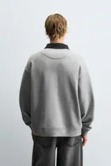 Sudadera gris de corte holgado, con cuello polo negro, cierre frontal de tres botones y mangas largas con puños acanalados.