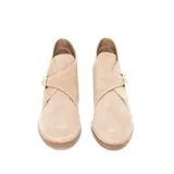 Bota corta de gamuza color avena, con diseño tipo botín chukka o desert boot modificado, que presenta una tira ancha cruzada sobre el empeine asegurada con una hebilla dorada. Posee taco bajo de madera.