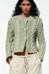 Chaqueta de punto color verde claro, con cuello redondo y manga larga. Cierre frontal con botones a presión en forma de flor.