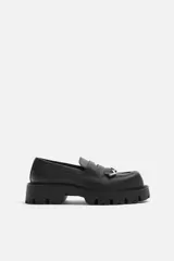 Mocasín negro de cuero con antifaz y flecos con puntas metálicas en el empeine, horma cuadrada y suela con volumen.