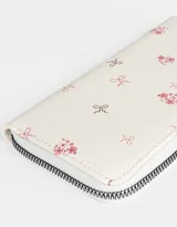 Billetera de cuerina color crema con estampado de flores y moños. Tiene formato rectangular, cierre circundante con tirador y múltiples compartimentos internos.