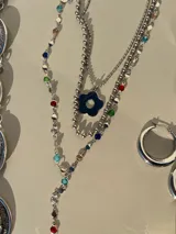 Set de tres collares plateados de diferentes largos. El más corto es tipo choker, el segundo tiene un dije de flor y el más largo es tipo corbatín con piedras de colores.