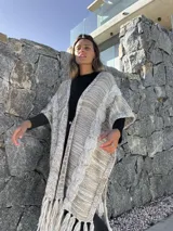 Ruana larga de lana merino color beige jaspeado, con diseño de trenzas y flecos en el borde inferior.