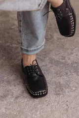 Mocasín de cuero negro con cordones y tachas metálicas en la puntera.