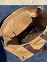 Bolso tote color beige con asas de cuero marrón, cierre superior y bolsillo interno.