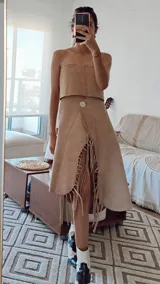 Conjunto color beige compuesto por un top strapless y una falda midi con abertura frontal y flecos. La falda tiene un botón decorativo en la cintura.