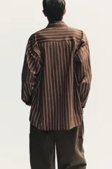 Camisa oversize marrón con rayas verticales blancas, cuello solapa, manga larga con puños y hombros caídos. Tiene un bolsillo de plastrón en el delantero, pinzas en la espalda y cierre frontal con botones.
