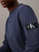 Buzo azul marino de algodón con cuello redondo y ribete en V. Tiene puños y dobladillo acanalados, y una insignia con el monograma de Calvin Klein en la manga.
