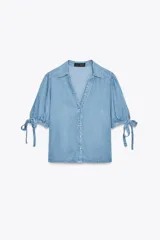 Camisa celeste de denim con cuello solapa, escote en pico, mangas abullonadas con lazos y cierre frontal con botones.