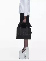 Bolso tote de cuero negro Marc Jacobs, tamaño mediano, con doble asa corta y correa larga extraíble. Estampado frontal con el texto "THE TOTE BAG" y el nombre de la marca.