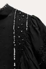 Blusa negra de viscosa con cuello perkins y manga larga. Detalles de bordados, lentejuelas y volantes. Cierre en la espalda con abertura y botones.