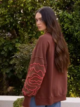 Buzo marrón de jogging sin friza con bordado floral en color coral en las mangas, corte holgado con tajos en los costados y puños.