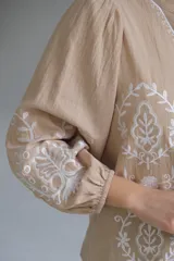 Camisa color beige con bordados florales blancos, cuello mao, mangas largas con elástico en los puños y cierre frontal con botones.