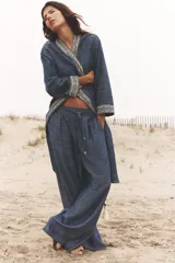 Pantalón ancho azul con textura de lino, tiro alto, cintura elástica con cordón ajustable, pinzas delanteras, bolsillos laterales y traseros, y cinturón de la misma tela con apliques metálicos.