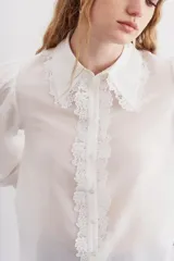 Camisa blanca de algodón con corte recto, cuello clásico y cierre frontal con botones. Presenta detalles de puntilla en cuello y cartera, mangas largas abullonadas con puños elastizados.