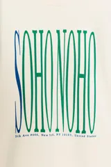 Sudadera de tejido mezcla de algodón color crema, con cuello redondo y manga larga. Presenta un estampado central con el texto "SOHO NOHO" en tipografía alargada, con las letras alternando entre azul y verde.