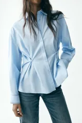 Camisa celeste de corte holgado, con cuello solapa, manga larga y cierre frontal con botones. Presenta detalle de cintura ajustable con botones.