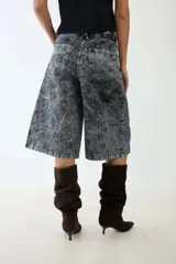 Bermuda de denim gris con acabado jaspeado y textura desgastada. Presenta un corte amplio y recto que cae por debajo de la rodilla, con bolsillos delanteros y traseros funcionales, trabillas para cinturón y cierre frontal con botón y cremallera.