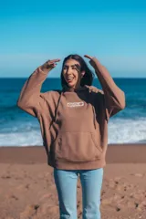 Canguro marrón con capucha y cordones ajustables. Presenta un estampado grande en la espalda con una ola, palmeras y el texto "Glassy Waves".