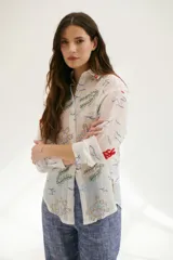 Camisa beige de manga larga con cuello clásico y cierre frontal con botones. Presenta bordados con motivos florales y geométricos en tonos azules, rojos y verdes.
