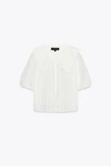 Camisa blanca de cuello bobo con bordados, escote pico con lazada, manga corta abullonada con elástico y cierre frontal con botones.