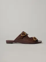 Sandalias planas de cuero color marrón con dos tiras anchas sobre el empeine, cada una con hebilla dorada.