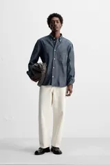 Camisa azul de algodón con textura chambray, corte regular fit, cuello abotonado, manga larga con puños abotonados, bolsillo de parche en el pecho y cierre frontal con botones.