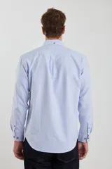 Camisa celeste de tejido texturado oxford, corte clásico, con cuello abotonado, mangas largas con puños ajustables, bolsillo en el pecho y cierre frontal con botones de madera.