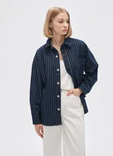 Campera tipo camisa de jean azul con rayas verticales blancas, corte holgado, cierre frontal con botones blancos y bolsillo de parche en el pecho.