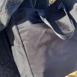 Bolso tipo tote color gris claro, con doble asa de mano y correa larga regulable. Tiene dos bolsillos laterales y un bolsillo frontal.