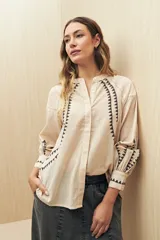 Camisa color crema con cuello mao y cierre frontal con botones. Presenta bordado con motivos geométricos en color negro en la parte superior del frente, mangas y puños.