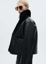 Chaqueta corta negra de cuero sintético con cuello y puños de piel sintética. Cierre frontal con botones y bolsillos de parche.