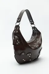 Cartera marrón de cuero sintético con forma de media luna, asa corta y apliques de tachas metálicas plateadas.