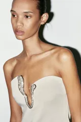 Vestido corto color crema, de escote asimétrico y sin mangas. Presenta una pieza metálica con apliques de strass en el escote.