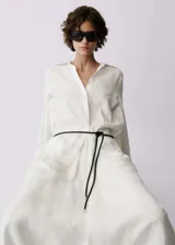 Camisa blanca de lino con cierre de botones en la parte delantera, mangas largas con puños abotonados, cuello camisero y corte recto. Incluye cinturón trenzado negro.