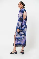 Vestido largo con estampado abstracto en tonos azules, violetas y grises. Tiene cuello mao con abertura en forma de gota, mangas largas acampanadas con detalle de botón en los puños, espalda abierta, tajo frontal y flecos en el ruedo.