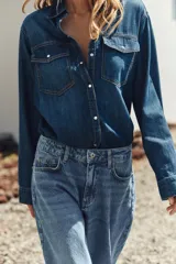 Camisa de denim azul con cuello solapa, manga larga, dos bolsillos delanteros con solapa y cierre frontal con botones a presión.