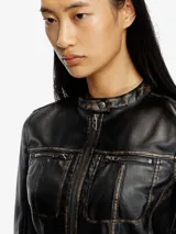 Chaqueta corta estilo biker para mujer, confeccionada en cuero sintético negro con detalles desgastados en las costuras. Presenta cuello mao con cierre a presión, cierre frontal con cremallera, bolsillos delanteros con cremallera y mangas largas.