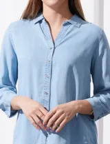 Camisa celeste de tencel con cuello con solapa, escote en V, abertura central con botones y mangas 3/4 regulables con presilla y botón.