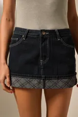 Minifalda de jean oscuro con costuras blancas contrastantes. El ruedo está doblado hacia afuera mostrando un forro o detalle de tela con estampado tartán en tonos grises y morados.