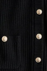 Cárdigan corto negro de punto acanalado, con cuello redondo, manga larga y botones metálicos dorados en el frente. Posee bolsillos de parche con botón en la parte inferior delantera.