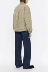 Cazadora corta color beige, de corte línea A, con cuello subido de punto, manga larga con puños fruncidos y cierre frontal con botonadura de presión. Tiene bolsillos con vivo y parche personalizado en el exterior.