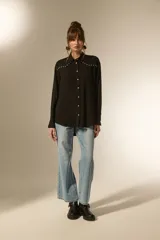 Camisa holgada de manga larga, color negro, con cuello camisa y botones frontales. Presenta apliques de tachas plateadas en los hombros y a lo largo de la tapeta frontal.