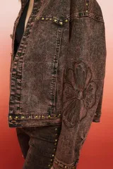 Campera de jean negra de corte holgado con detalles de tachas plateadas en costuras, hombros y puños, y bordado floral blanco en las mangas.