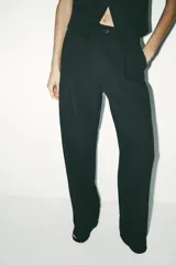Pantalón de tiro alto color negro, confeccionado en hilatura con 22% de lino. Presenta bolsillos delanteros, detalle de pliegues, y cierre frontal con cremallera y botón.
