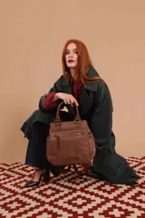 Mochila bandolera color marrón con herrajes color peltre. Tiene un compartimento principal amplio con cierre, un bolsillo frontal con cierre y dos bolsillos laterales abiertos. Incluye correa ajustable para usar al hombro o cruzada.