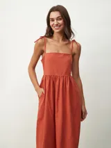 Vestido midi de color naranja, con escote recto y tirantes finos que se anudan sobre los hombros. Posee una cintura entallada con frunces que dan paso a una falda fluida.