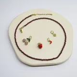 Gargantilla de cuentas facetadas color borgoña con cierre de mosquetón dorado y cadena de alargue. Incluye cuatro dijes colgantes dorados: cerezas con strass rosa, frutilla roja, corazón rojo esmaltado y estrella con strass.