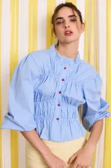 Camisa celeste a rayas verticales blancas, de corte entallado, con cuello clásico, cierre frontal con botones rojos, frunces en el delantero y mangas largas abullonadas con puños elásticos.